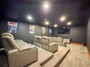Sala de cine en casa con varios sillones de cuero color beige, un televisor de pantalla plana y carteles de películas en la pared.