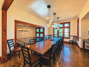 A spacious dining area with a long wooden table surrounded by chairs, large windows, and a chandelier.