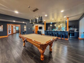 Una habitación amplia con mesa de billar, un área de bar con taburetes y una cocina al fondo.