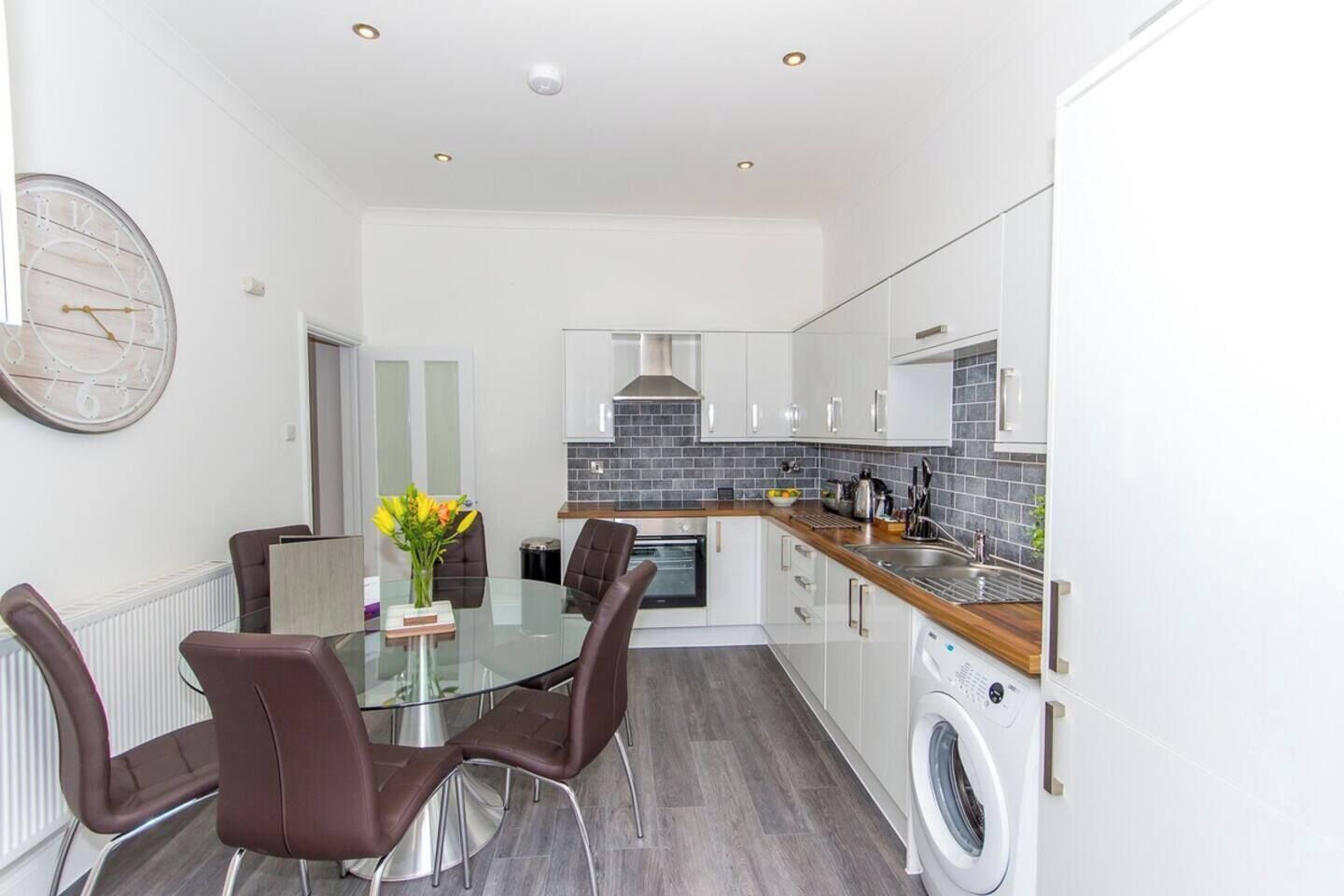 A modern kitchen with a glass dining table, brown chairs, and a built-in washing machine.
