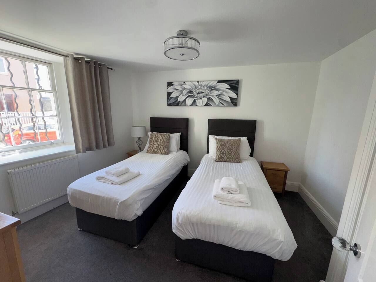 Ein Hotelzimmer mit zwei Einzelbetten, einem Vorhangfenster, einem Heizkörper, einem Wandbild und einer Deckenleuchte.