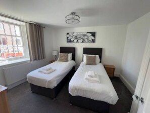 Habitación de hotel con dos camas individuales, una ventana con cortinas, un radiador, una obra de arte en la pared y una lámpara en el techo.