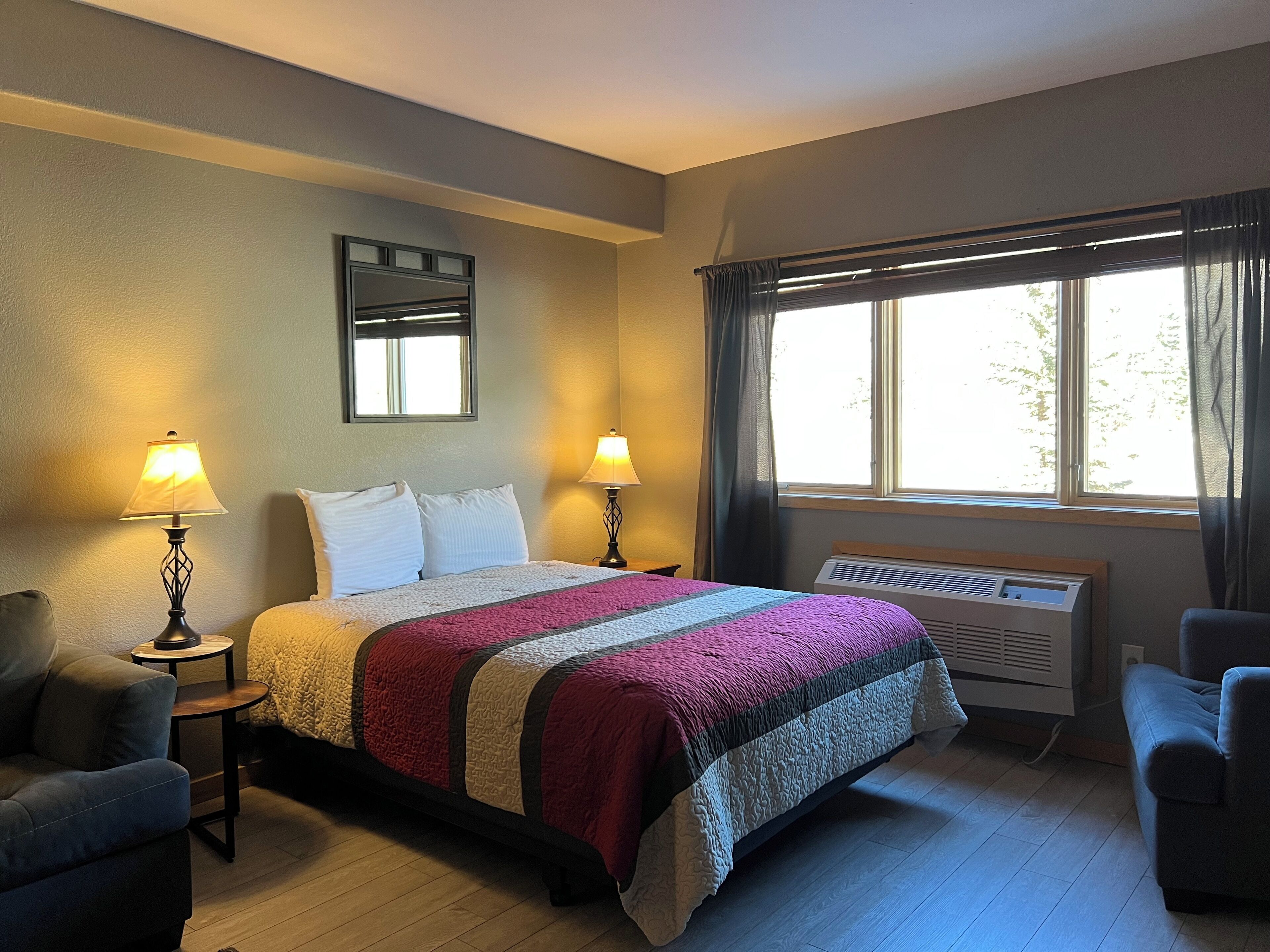 A hotel room with a bed, two bedside lamps, a window with curtains, a radiator, and a small table with a chair.