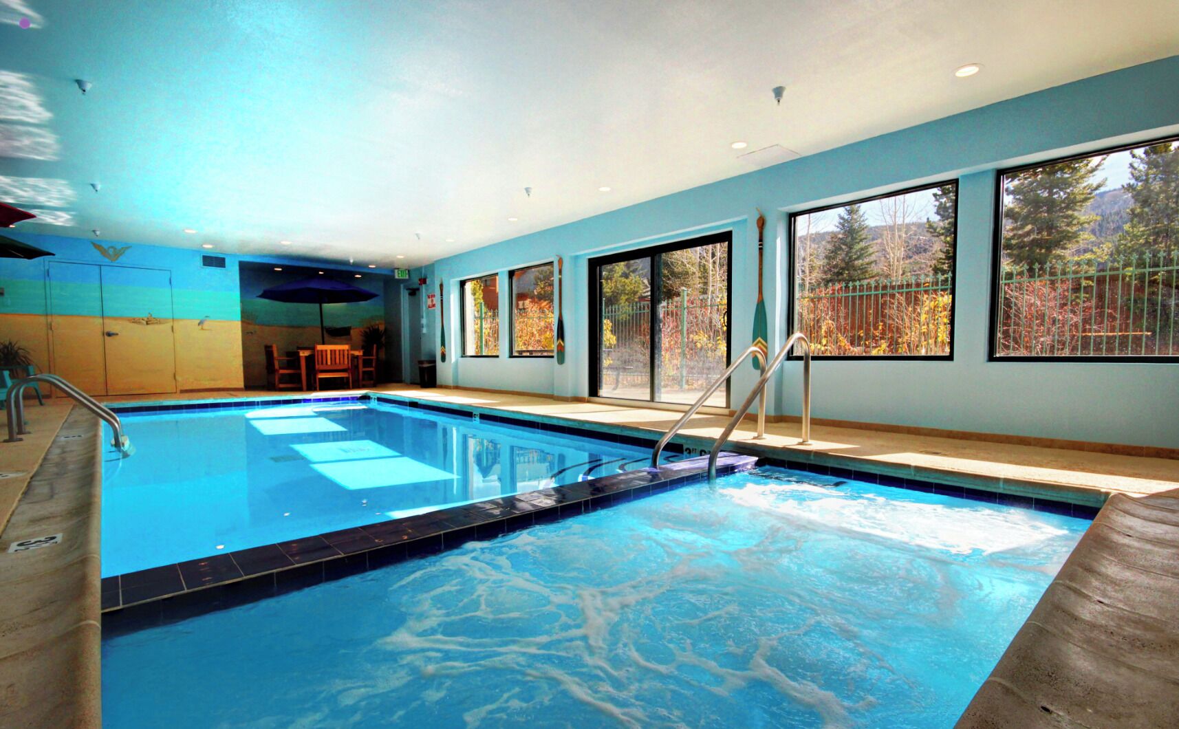 Piscina interior com água cristalina, deck de madeira e grandes janelas com vista para árvores e montanhas.
