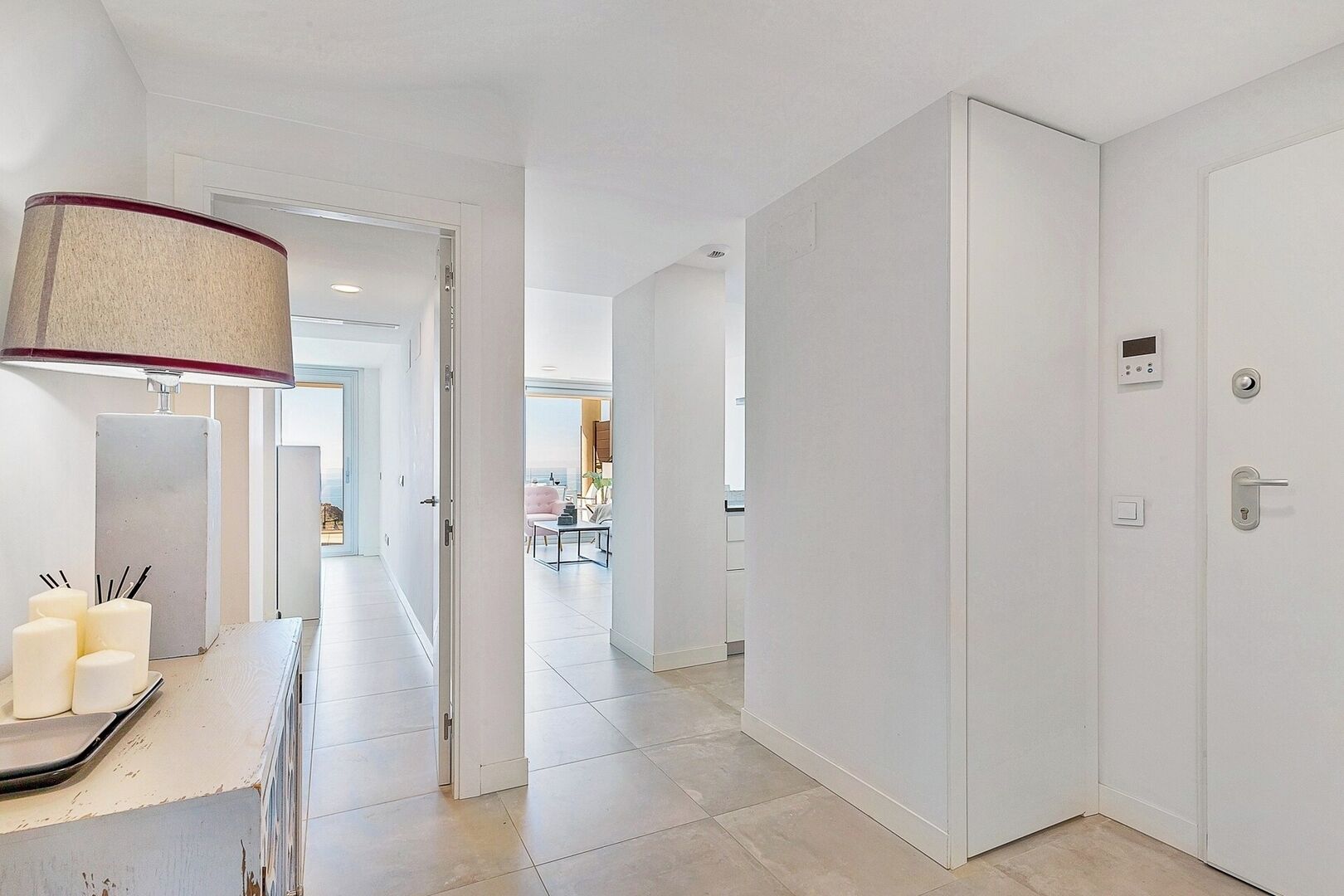 A modern hallway with a lamp, a door with a digital lock, and a view into a living area with a dining table and chairs.