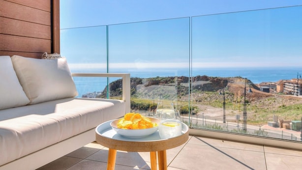 Een balkon met een witte bank, een kleine ronde tafel met een schaal chips en een glas water, en een helder uitzicht op de kustlijn.