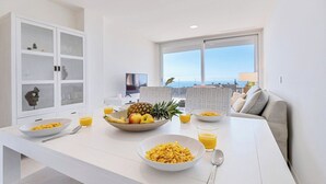 Een moderne keuken met witte kastjes, een ontbijt gedekte eettafel en uitzicht op de oceaan door grote ramen.