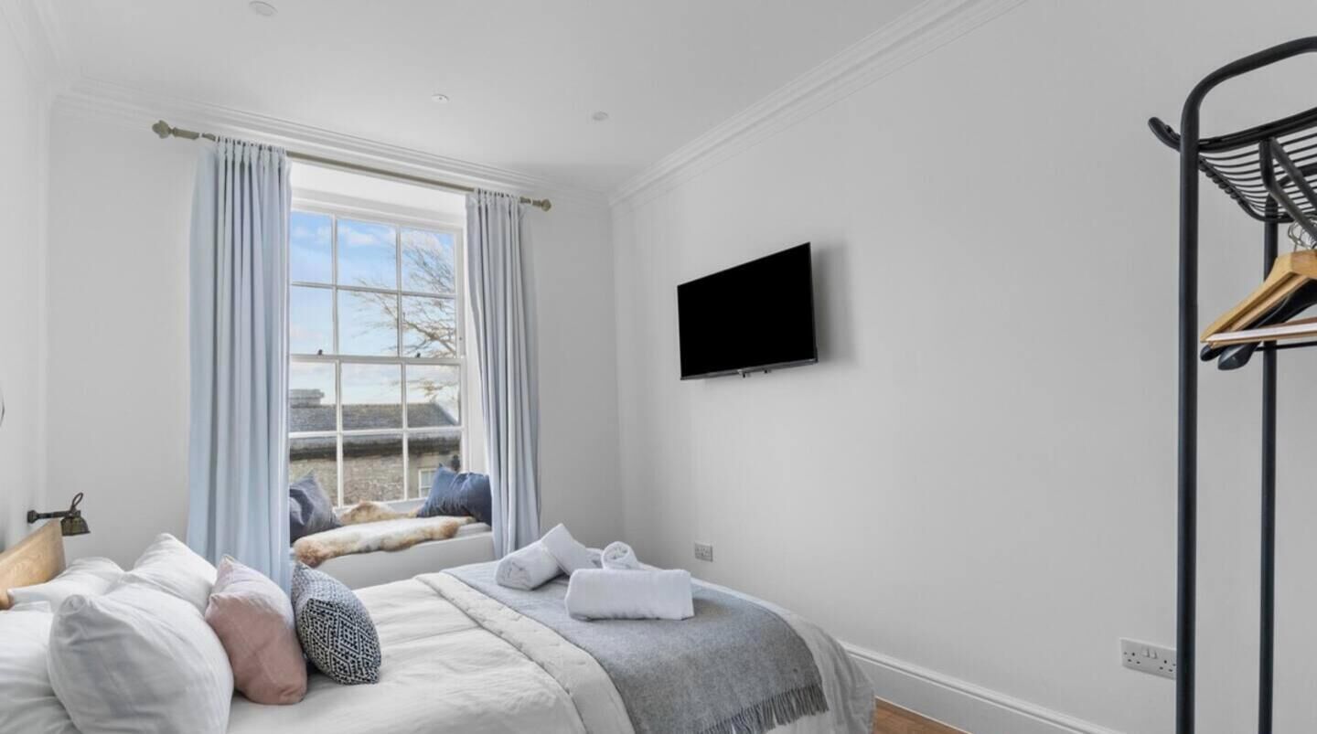A hotel room with a bed, a window with curtains, a television on the wall, and a coat rack.