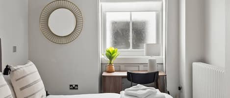 Une chambre à coucher comprenant un lit, un bureau, une chaise, une plante en pot et une fenêtre avec des stores.