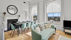 A modern living room with a fireplace, dining area, and large windows offering a view. - Hurlingham by Pureserviced (Plymouth)