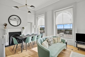 A modern living room with a fireplace, dining area, and large windows offering a view. - Hurlingham by Pureserviced (Plymouth)