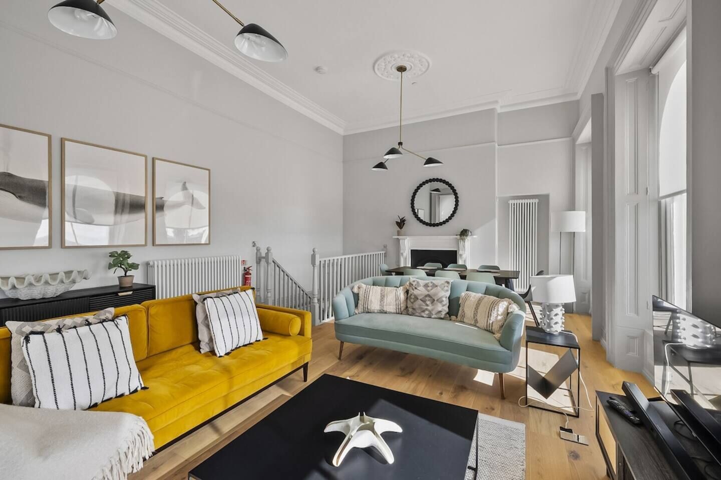Un salon moderne avec un canapé jaune, un canapé turquoise et une table basse noire. On y trouve des tableaux encadrés au mur, une cheminée et un escalier.