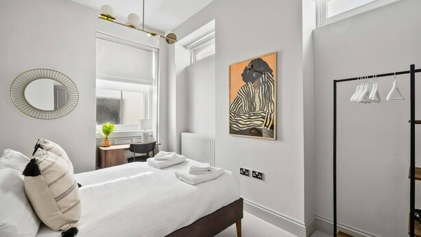 A modern bedroom with a bed, bedside table, mirror, artwork, and a clothing rack. - Hurlingham by Pureserviced (Plymouth)