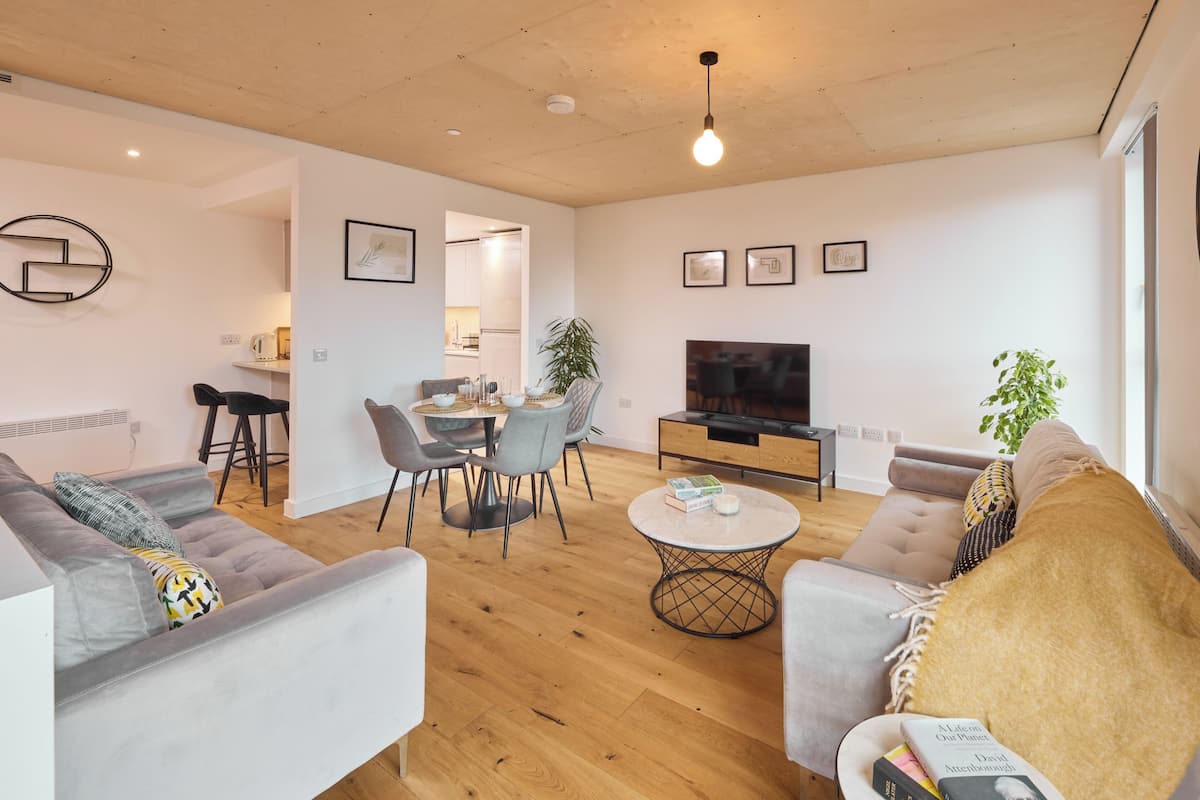A modern living room with a sofa, coffee table, and TV. There is a dining area with a table and chairs. The room has wooden flooring and white walls.