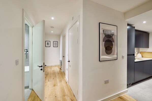 A modern hallway with wooden flooring, white walls, and a kitchen area with dark cabinets and a white countertop. - Finlays Yard by Veeve (Manchester)