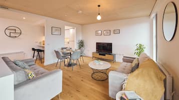 A modern living room with a sofa, coffee table, and TV. There is a dining area with a table and chairs. The room has wooden flooring and white walls.