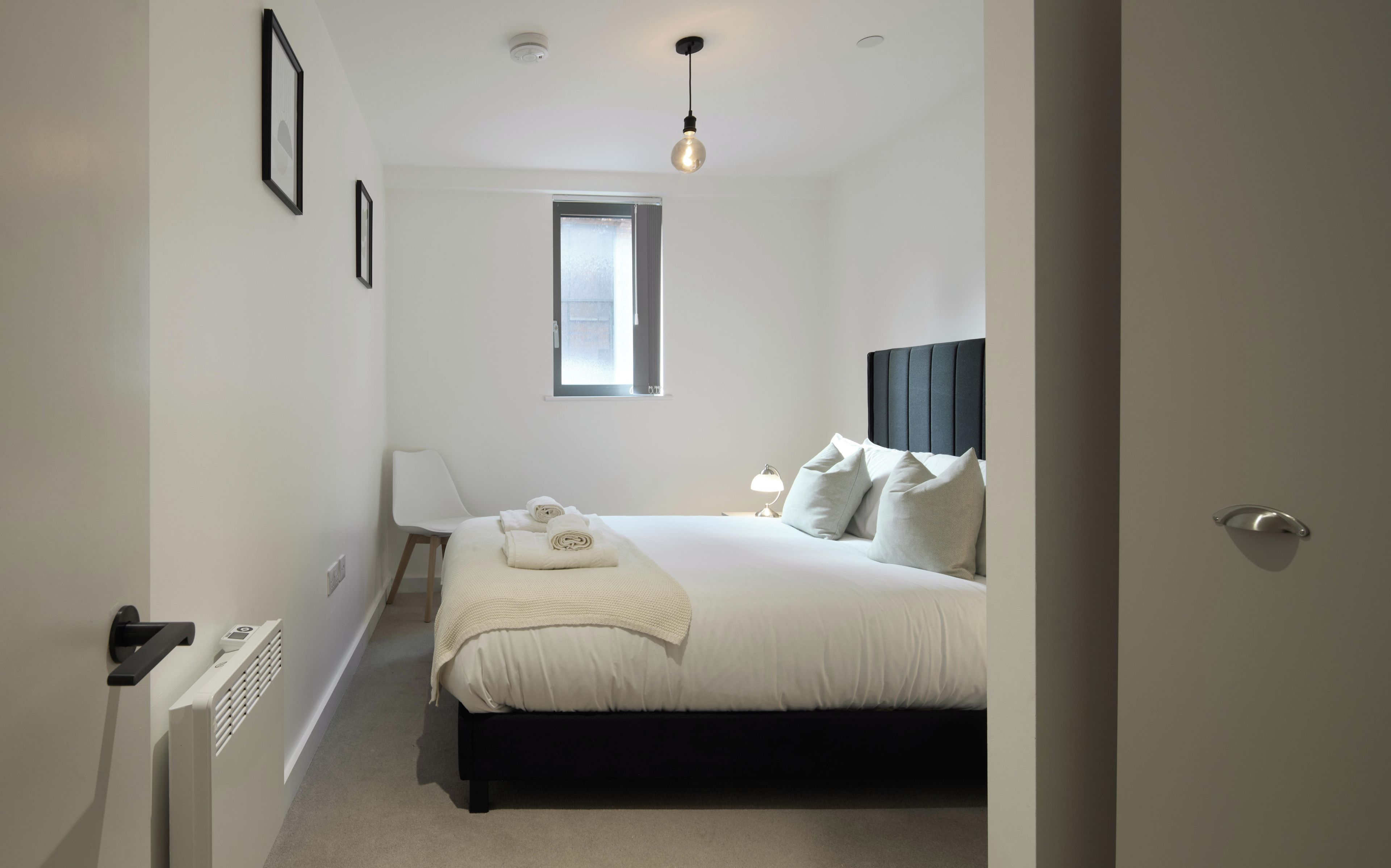A bedroom with a bed, a window, a radiator, a chair, and a bedside table.