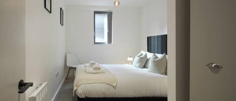 A bedroom with a bed, a window, a radiator, a chair, and a bedside table.