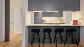 Una cocina moderna con encimera blanca, horno empotrado, un lavabo y una cafetera roja. Hay cuatro taburetes negros debajo de la encimera.