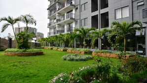 Un moderno edificio de apartamentos con varios balcones, un jardín cuidado con palmeras y macizos de flores, y una vista despejada del entorno urbano.