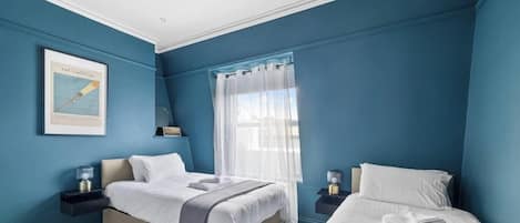Un dormitorio con dos camas, un ventanal con cortinas translúcidas, un cuadro en la pared y lámparas de noche.