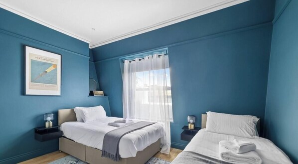 A bedroom with two beds, a large window with sheer curtains, a framed picture on the wall, and bedside lamps.
