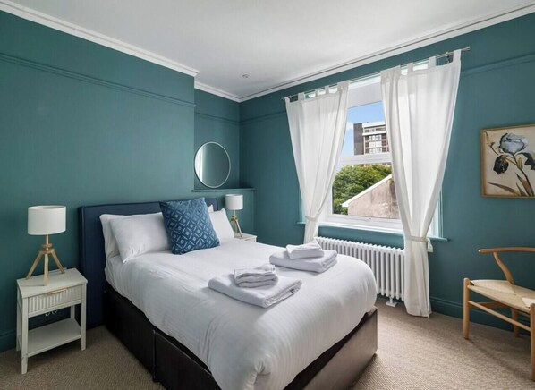 A bedroom with a bed, a nightstand, a chair, a window with curtains, and a painting on the wall.