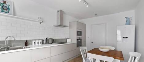 Una cocina moderna con armarios blancos, un lavamanos de acero inoxidable y una mesa de comedor de madera con sillas blancas.