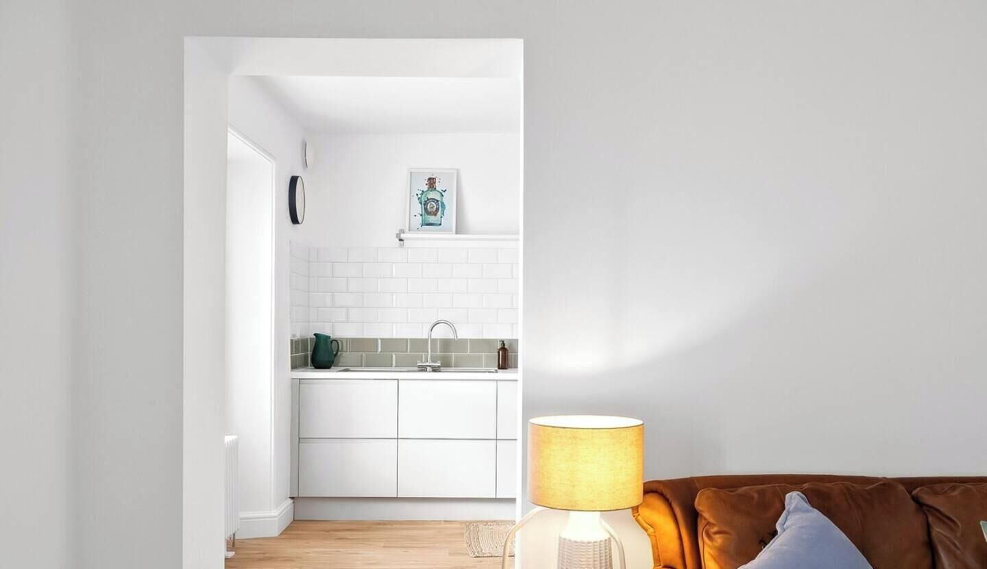 Una cocina moderna con armarios blancos, salpicadero de azulejos, un lavamanos y una encimera con una tetera y una botella.