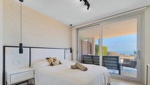 A modern bedroom with a large bed, a nightstand, a bedside table, and a view of the outdoors through a sliding glass door. - Modern 3bdr aprt - Ocean view (Fuengirola)