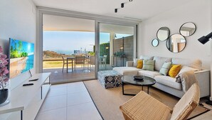 A modern living room with a large TV, a sofa, a coffee table, and a view of the beach. - Modern 3bdr aprt - Ocean view (Fuengirola)