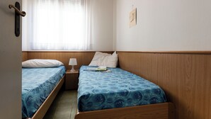 A small, neatly arranged room with two single beds, a bedside table with a lamp, a window with sheer curtains, and a wall-mounted picture. - Cozy Apartment Near the Beach in Bibione (San Michele al Tagliamento)