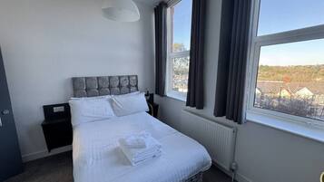 Un dormitorio con cama, mesita de noche, ventana con cortinas y vistas al exterior.