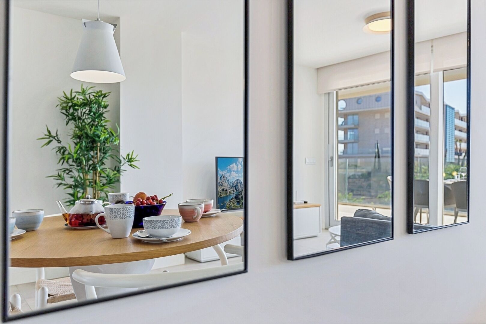 A modern dining area with a wooden table, white chairs, and a large mirror reflecting the room. There is a potted plant and a framed picture on the table.