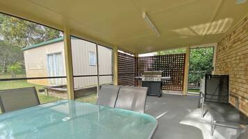 Une terrasse couverte avec une table et des chaises en verre, un barbecue et une vue sur le jardin arrière.