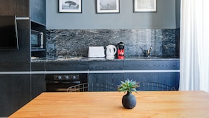 A modern kitchen with dark cabinetry, a marble backsplash, and a dining area with a wooden table and chairs. There are framed pictures on the wall above the counter. - Moderno Studio en Palermo Hollywood C3D (Buenos Aires)