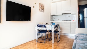 A compact living space with a TV mounted on the wall, a dining table with chairs, a kitchenette, and a bed. - Great Apartment With Sunny Terrace in Bibione (San Michele al Tagliamento)