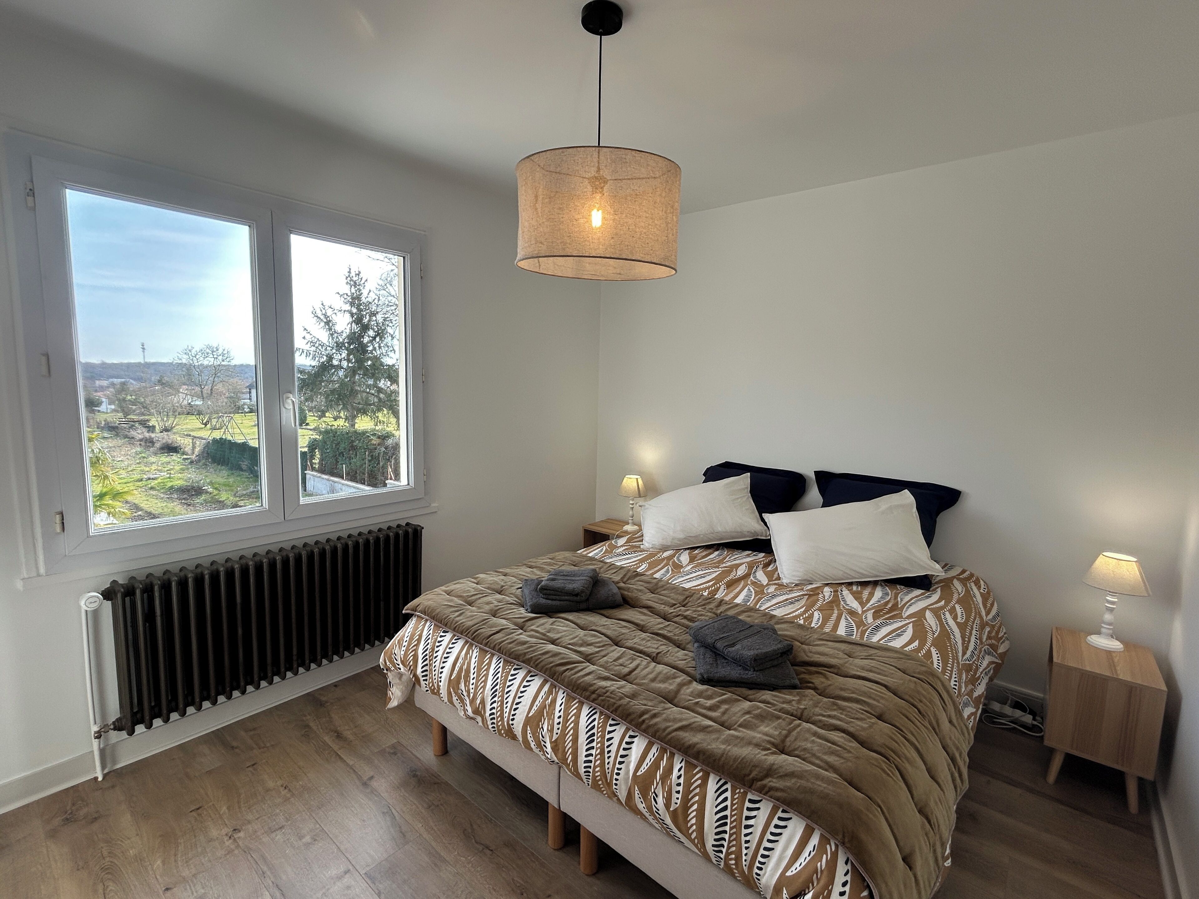 A bedroom with a large bed, a window with a view, a radiator, and a bedside table with a lamp.