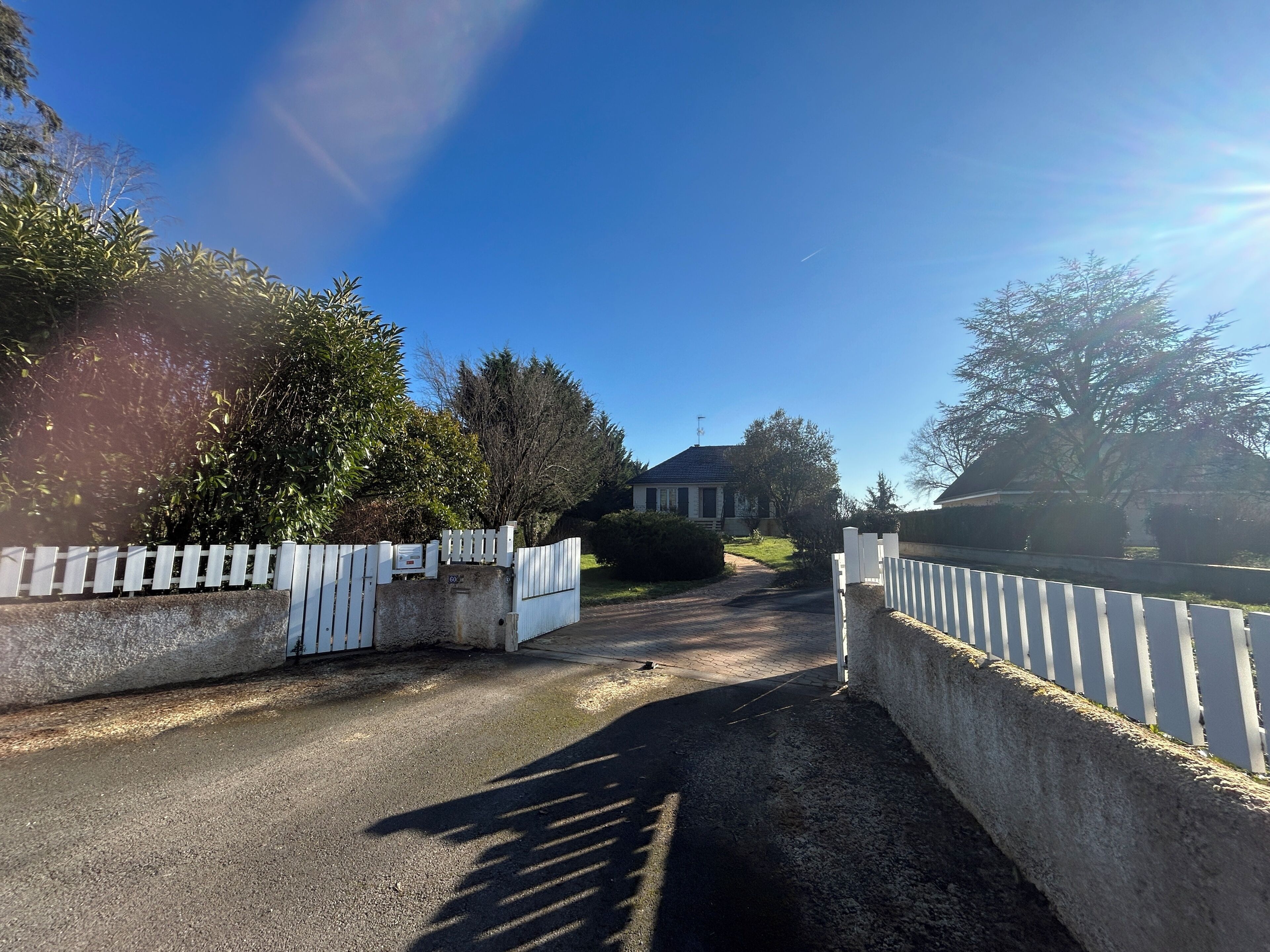 Una calle residencial con una entrada porticada, una casa al fondo y un cielo azul despejado.