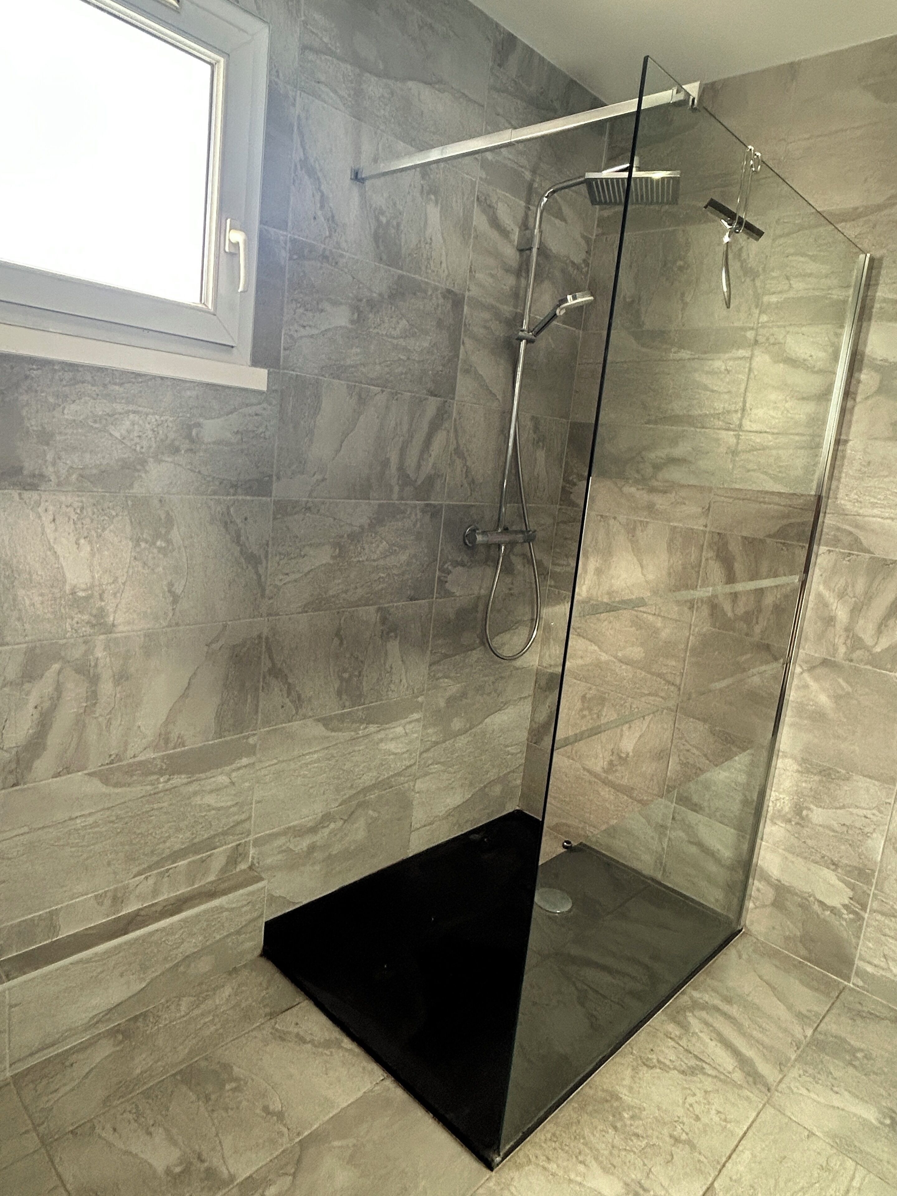 Baño con ducha envuelta en vidrio, piso de felpudo negro, paredes de mármol gris y una ventana arriba.