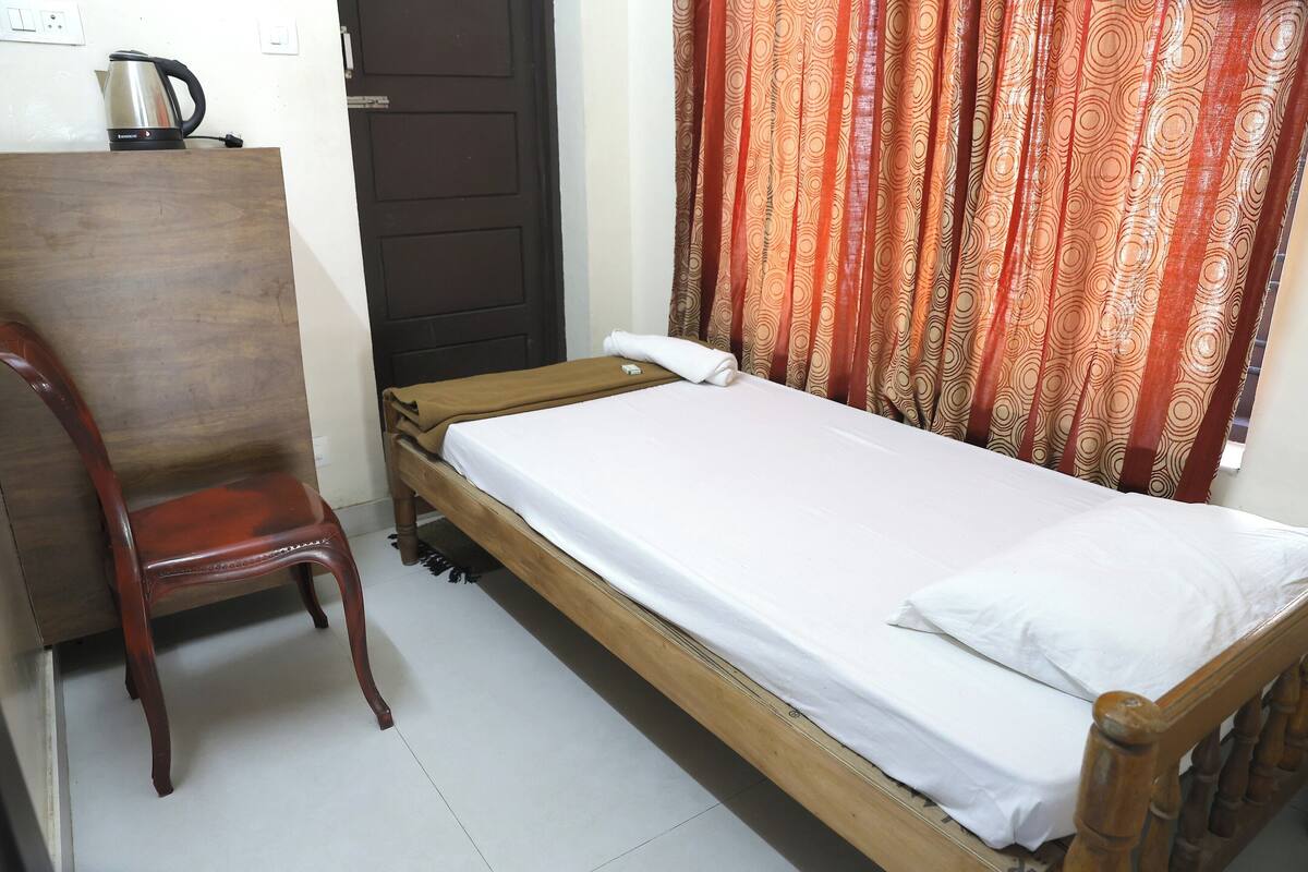 A single bed with a wooden frame and white bedding, a wooden chair, a kettle on a small table, and a door with a curtain.