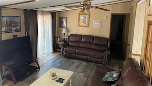 Una sala de estar con un sofá de cuero marrón, un televisor, un ventilador de techo y una mesa de centro con controles remotos.