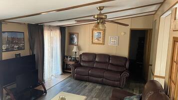 A living room with a brown leather sofa, a television, a ceiling fan, and a coffee table with remotes.