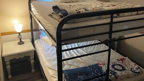 Una cama litera con una mesita de noche y una lámpara, una ventana con persianas, y suelo de madera.