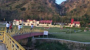 A hotel with multiple red-roofed buildings, a grassy lawn, and a bridge leading to the entrance. - Tikkar Taal Morni Hills (Panchkula)