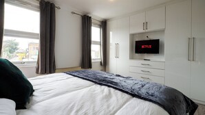 A hotel room with a bed, a TV mounted on the wall, and a window with curtains. - Dartford - 3 Bed House- Sleeps7 - Garden (Dartford)
