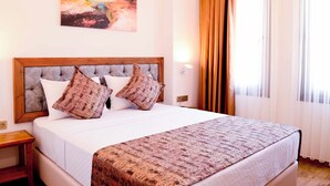 A neatly made bed with a patterned bedspread and two decorative pillows, a painting on the wall, and a window with curtains. - Troiada Hotel (Gokceada)