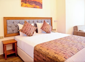 A neatly made bed with a patterned bedspread and two decorative pillows, a wooden nightstand, and a chair in a well-lit room with a simple wall art piece above the bed. - Troiada Hotel (Gokceada)