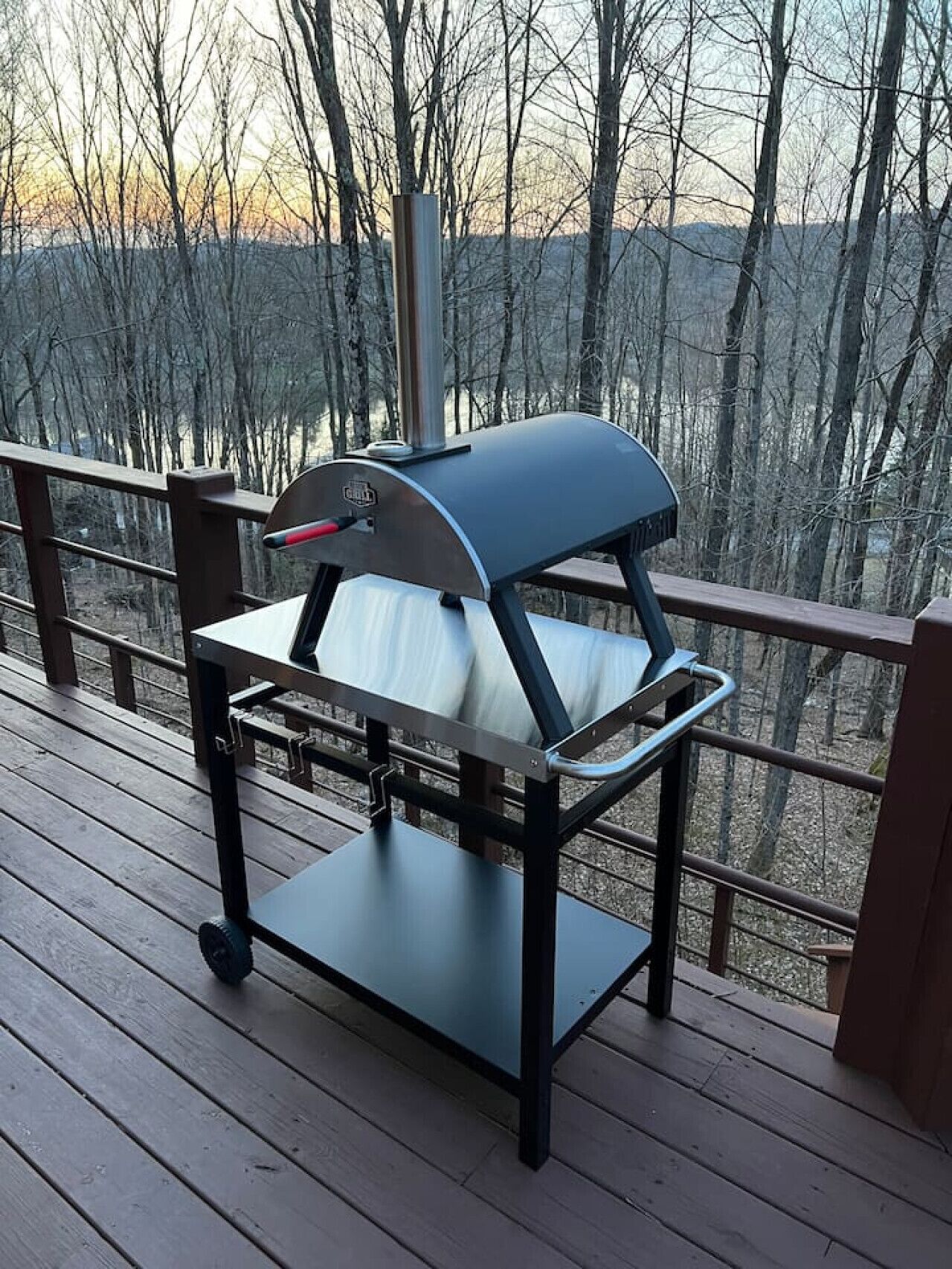 A portable grill on a wooden deck with a railing, overlooking a forested area.