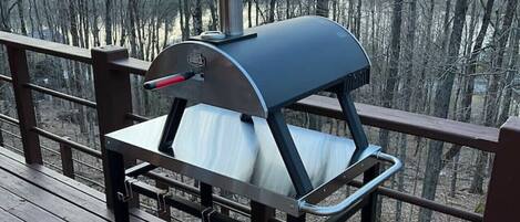 A portable grill on a wooden deck with a railing, overlooking a forested area.